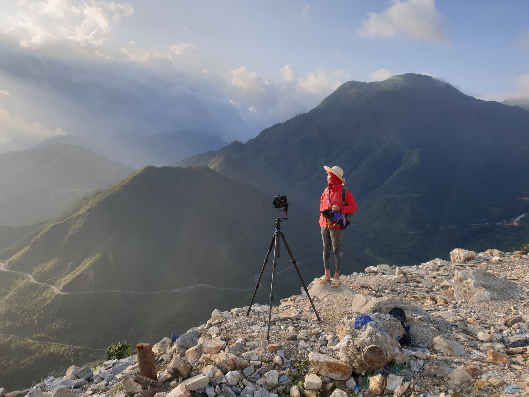 person wearing red jacket standing on cliff beside black manfrotto befree camera tripod