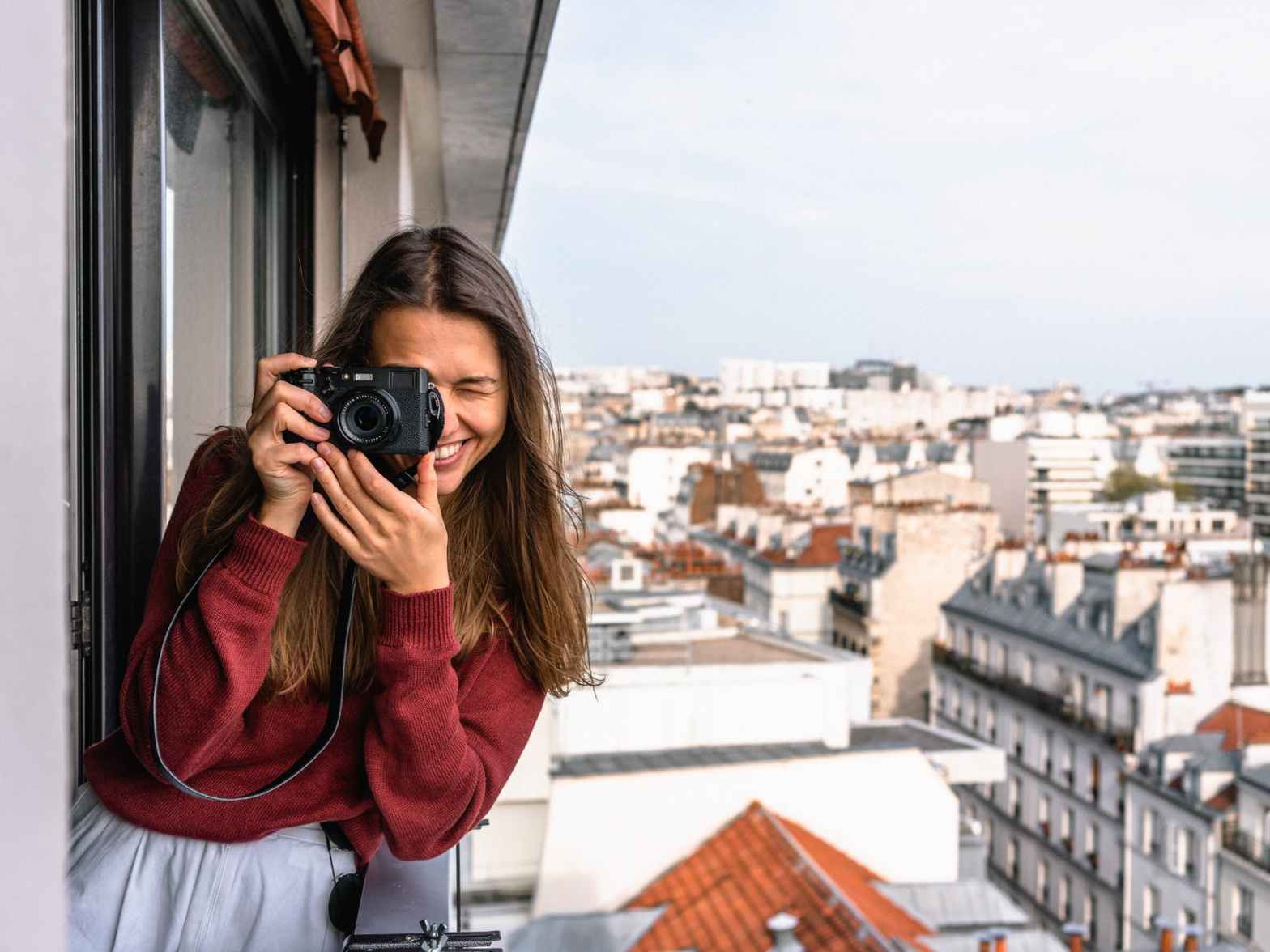woman wearing maroon sweater standing on veranda using camera while smiling overlooking houses and buildings